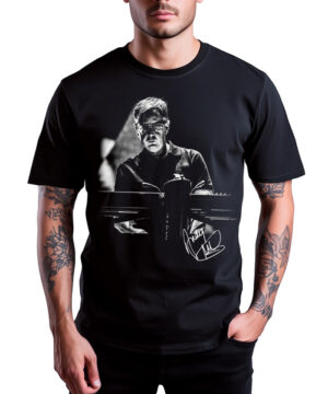 Osoba nosząca DEPECHE MODE "Fletch" - czarną koszulkę z nadrukowanym wizerunkiem mężczyzny grającego na keyboardzie i białą grafiką z podpisem.