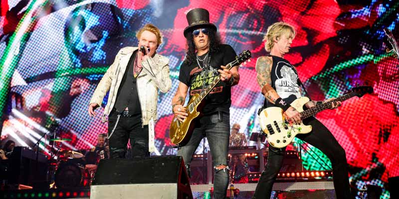 Czarna koszulka Guns N' Roses "Think About You" noszona przez basistę podczas koncertu rockowego, scena z kolorowymi światłami.